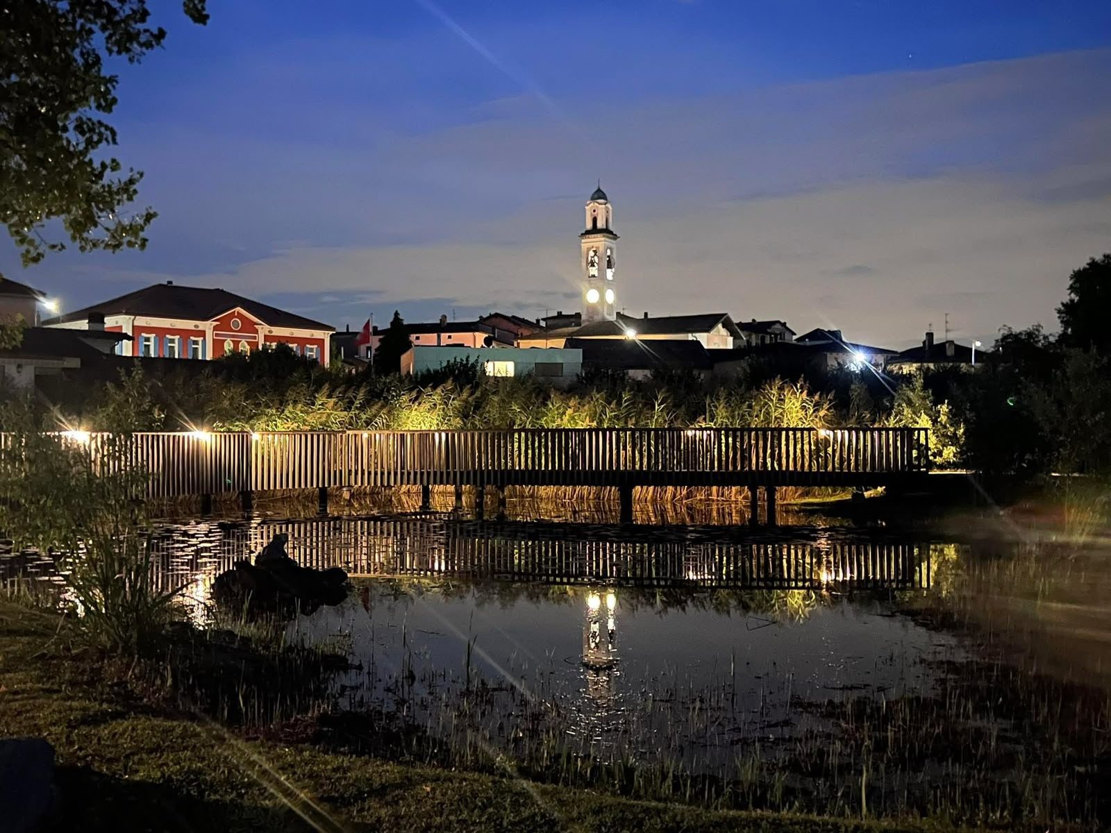 Il laghetto di Coldrerio illuminato di sera, con la passerella di legno e il campanile riflesso nell'acqua